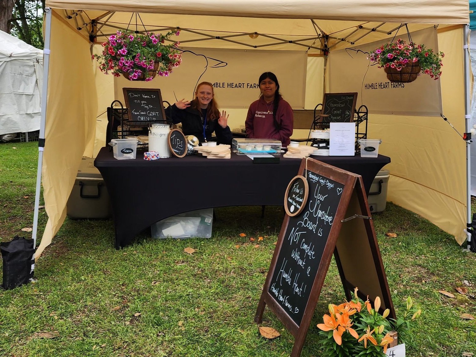 Farmers market booth for Humble Heart Farms featuring two smiling vendors. The table displays various cheese products and a chalkboard sign promoting their offerings. Colorful hanging flowers add to the vibrant atmosphere.