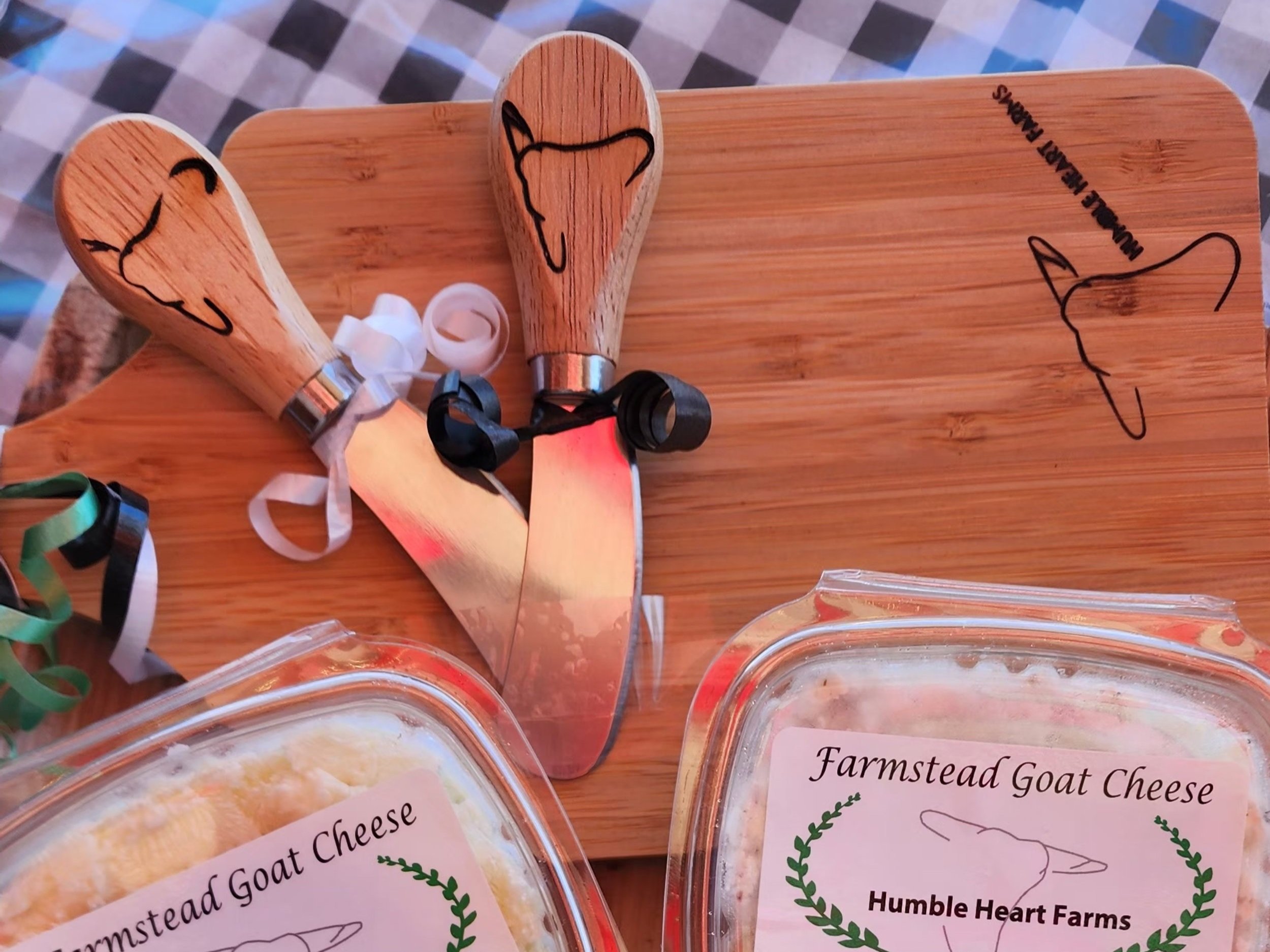 Farmstead goat cheese from Humble Heart Farms displayed with cheese knives on a wooden cutting board. The image features artisan cheese packaging and decorative ribbons, highlighting a farm-fresh culinary experience.