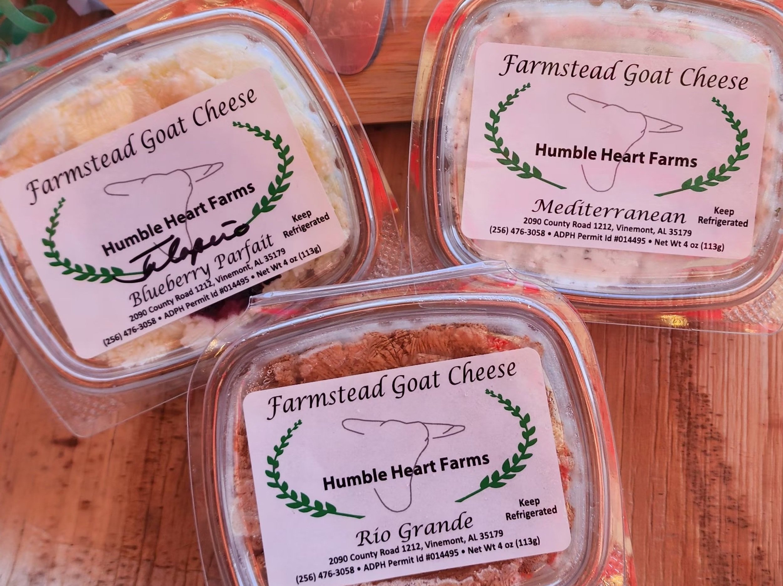 Farmstead goat cheese varieties from Humble Heart Farms, including Blueberry Parfait, Mediterranean, and Rio Grande, displayed in clear containers on a wooden surface. Each label includes storage instructions and contact information for the farm.