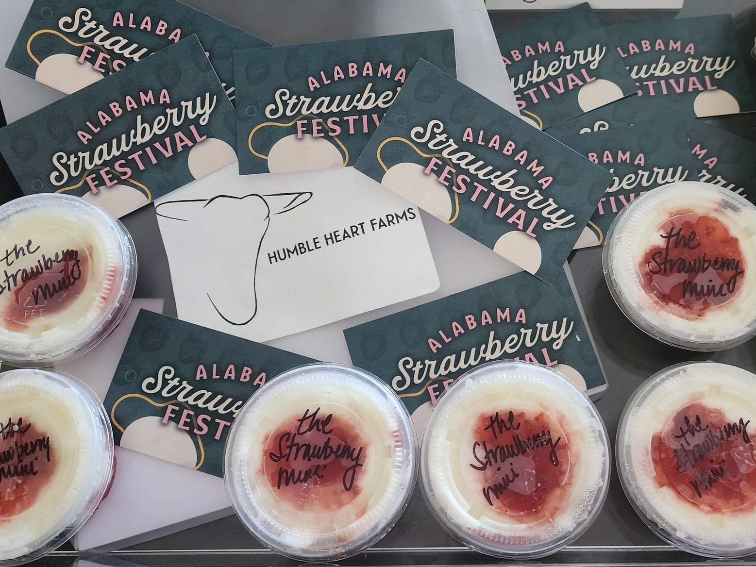 Cups of strawberry dessert labeled "the Strawberry Mini" alongside promotional cards for the Alabama Strawberry Festival and a business card for Humble Heart Farms, showcasing local food and festival culture.